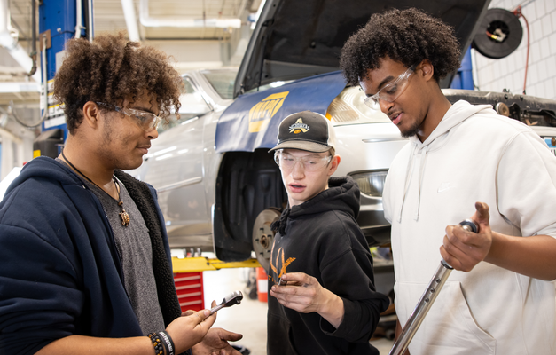 Three individuals discussing tools in an automotive workshop. A car is visible in the background on a lift.