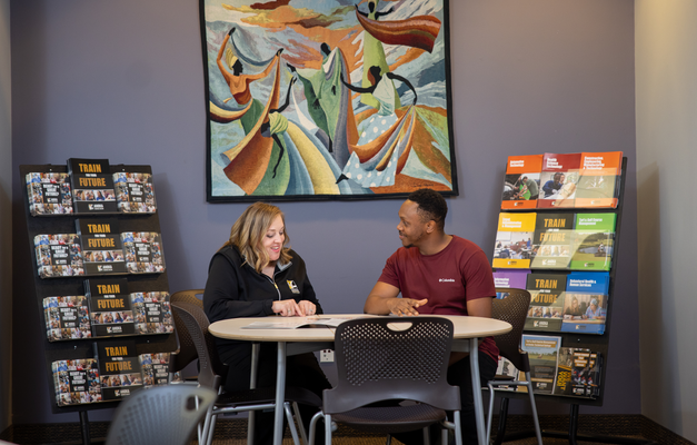 Two people are seated at a table in a room with informational brochures and a vibrant mural on the wall. 