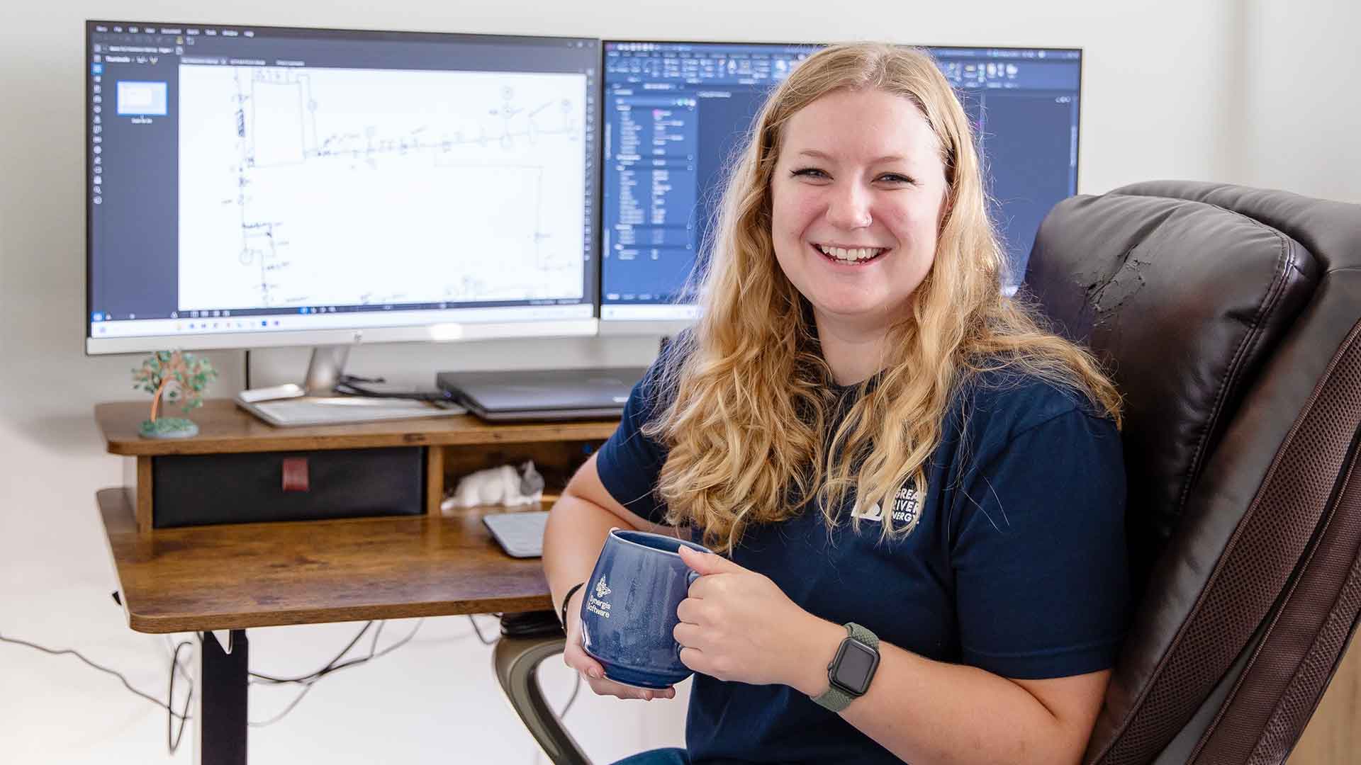 Individual smiling at the camera, holding a mug with a dual monitor desktop setup displaying blueprints in the background.