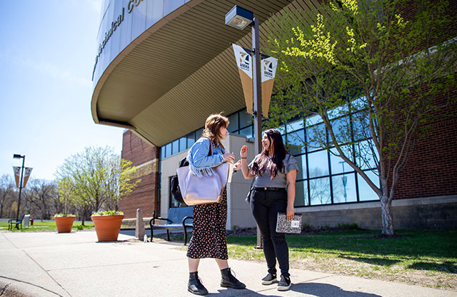 two female students talking outside with college building entrance in background