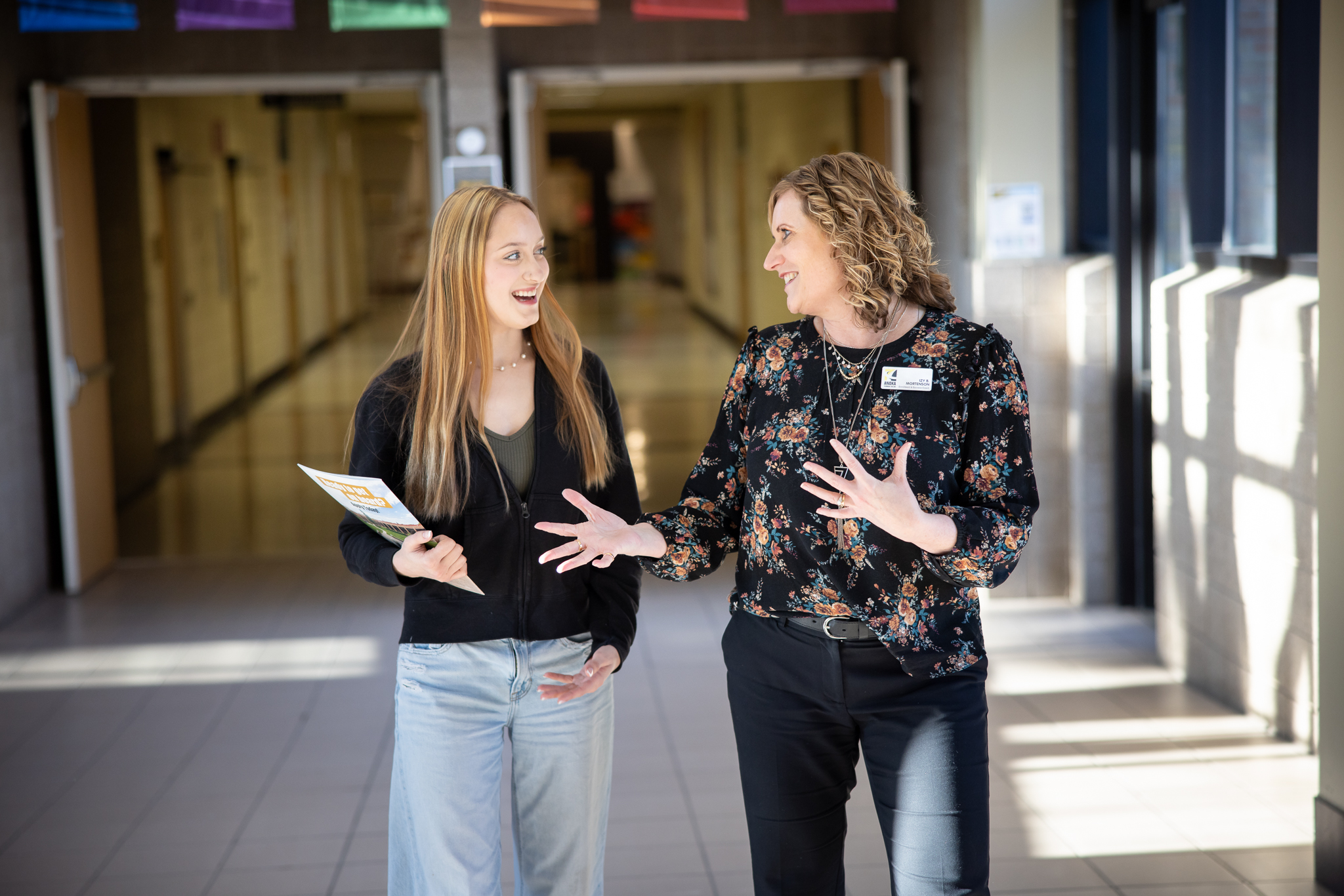 Two individuals are having a conversation in a brightly lit school hallway, one holding papers. They appear engaged and happy, standing near classrooms.